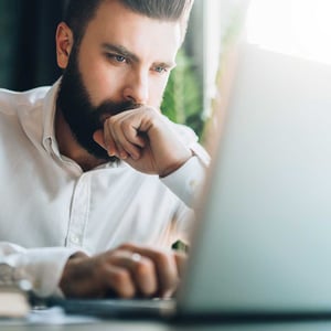 young-serious-bearded-businessman-sitting-in-office-at-table-and-using-laptop.